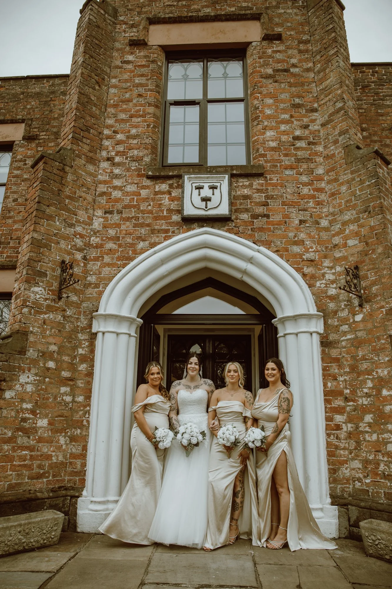 Bride and bridesmaids in beige dresses holding bouquets outside a brick building with an arched entrance.