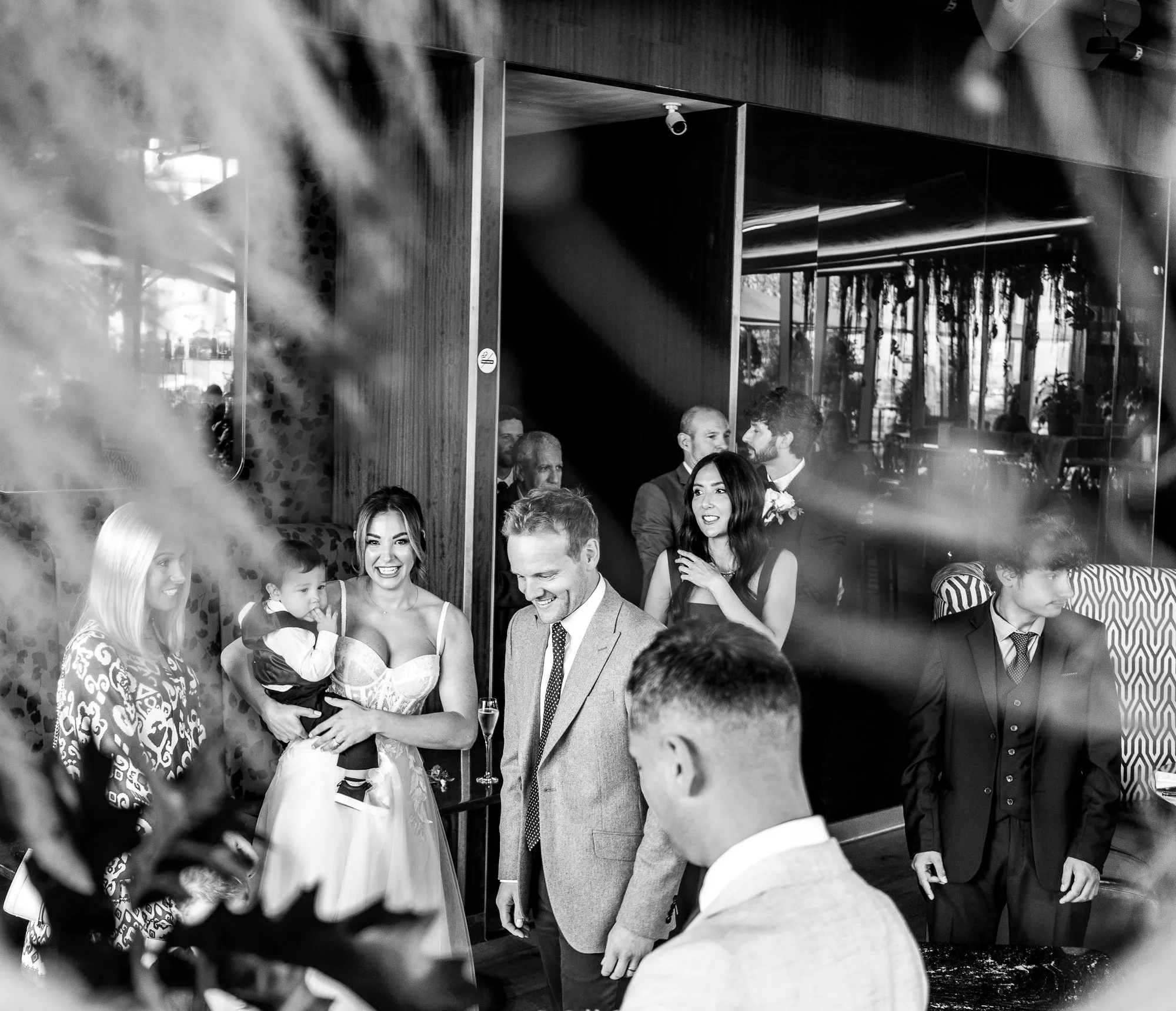 A black and white photo of people gathered indoors, with a woman in a wedding dress holding a child. Several other people are dressed in formal attire, likely at a wedding or celebration. Reflections and plants partially obscure some of the scene.