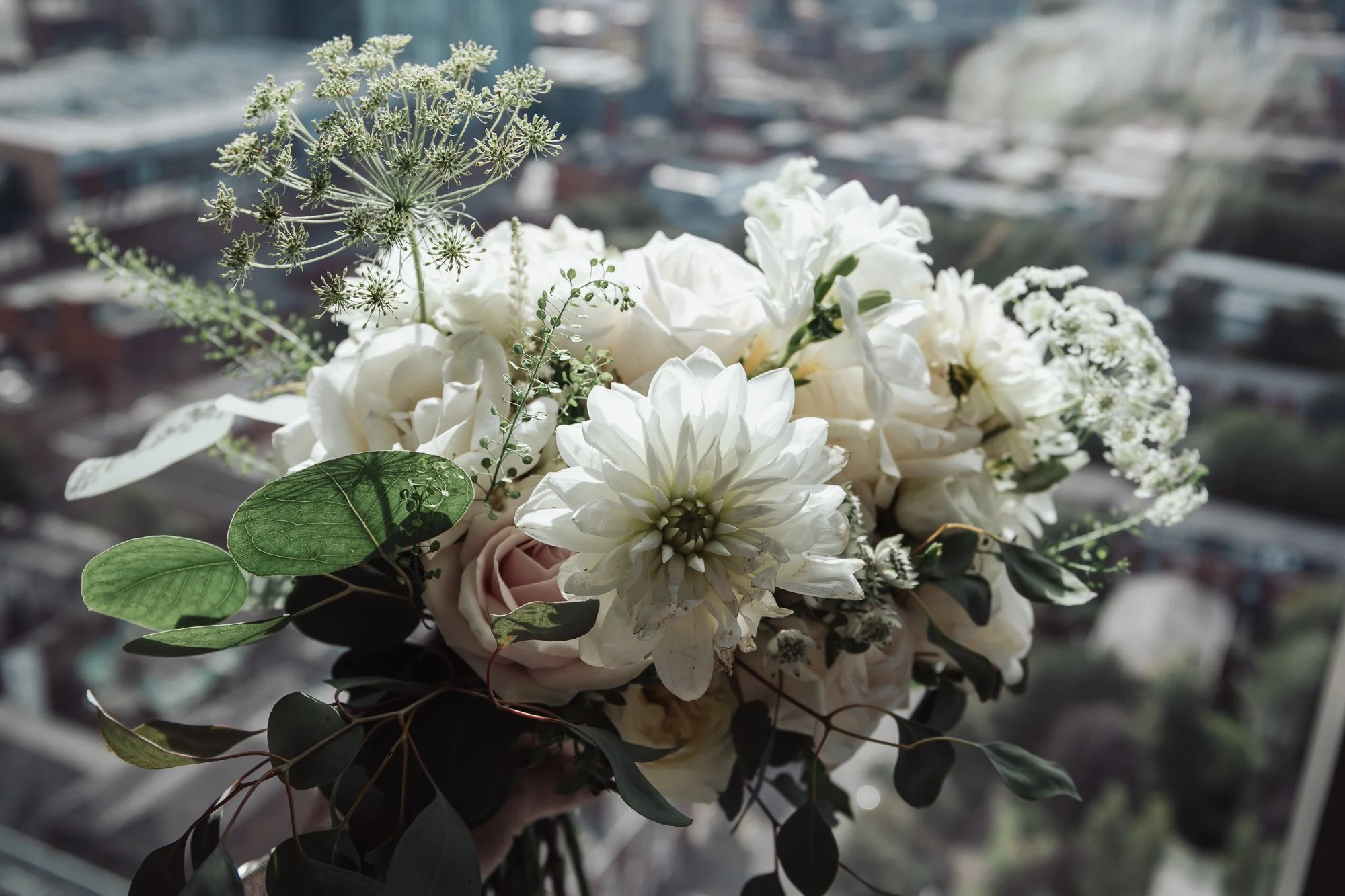 Bouquet of white flowers with greenery, blurred cityscape in background.