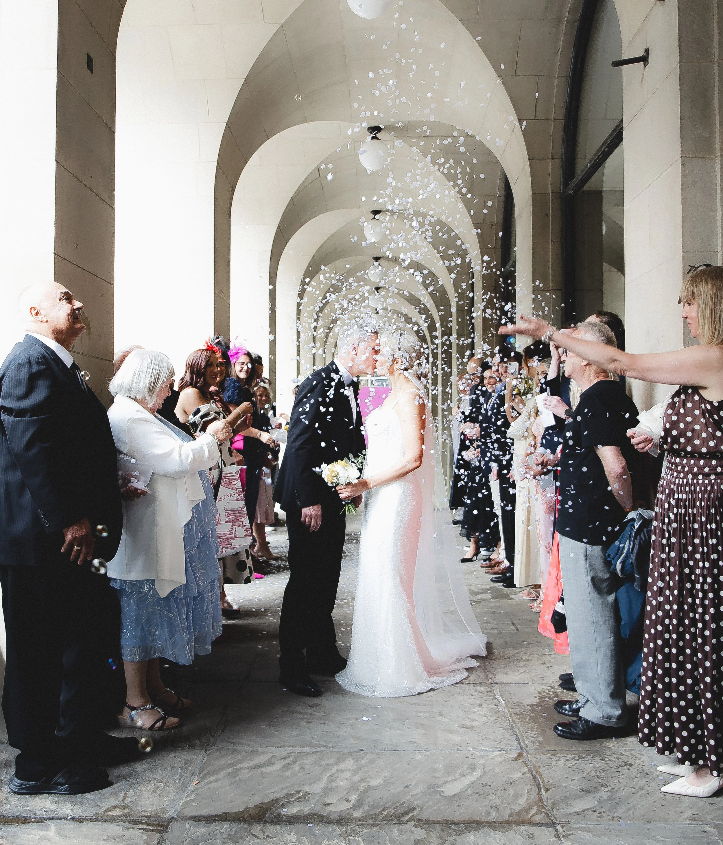 A wedding celebration with guests standing under an archway, throwing confetti, and watching a bride and groom kiss. The guests are dressed in formal attire, and the setting appears to be outdoors with a stone or concrete floor and a series of archways in the background.