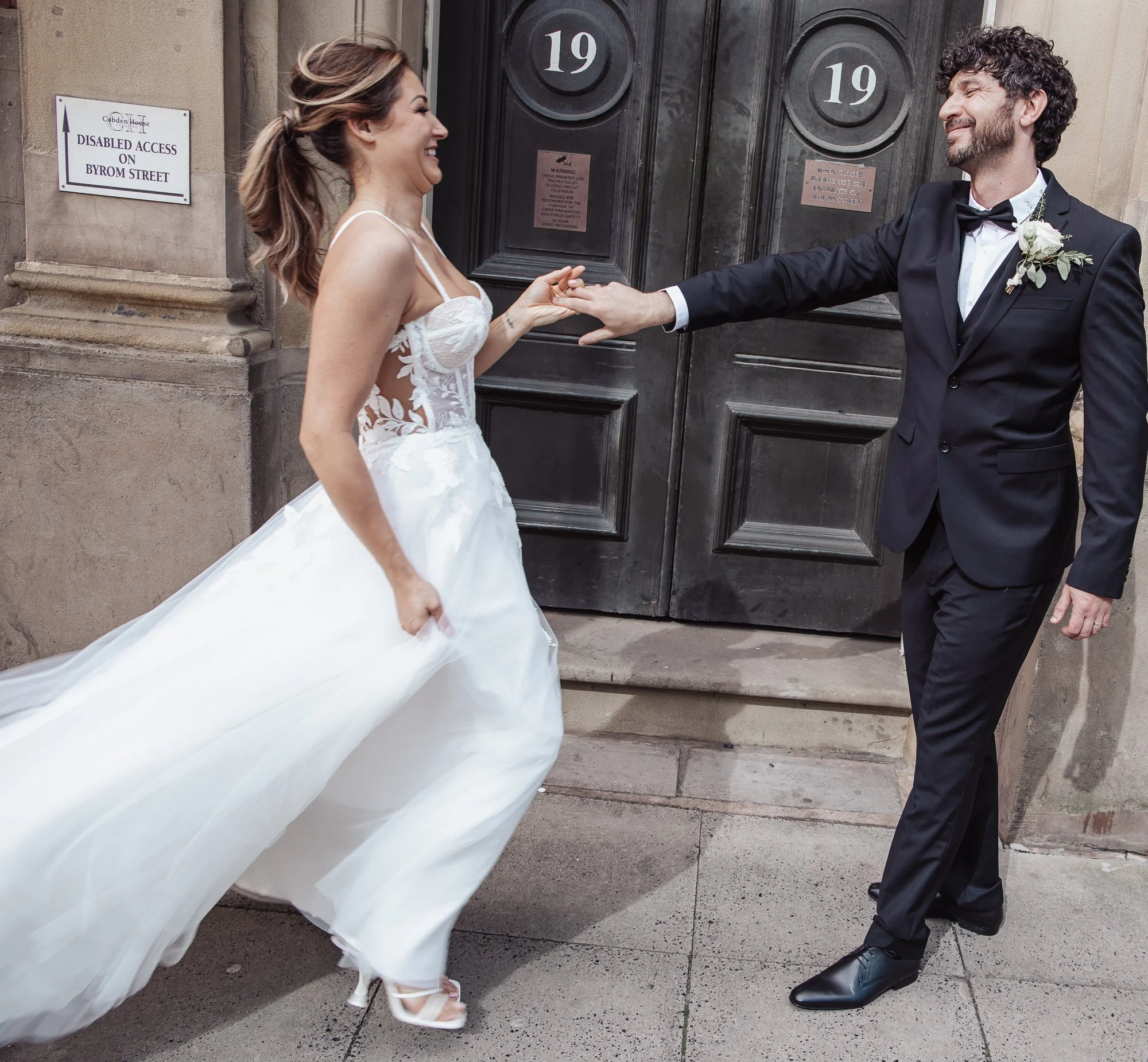 A bride in a white wedding dress and a groom in a black suit are holding hands and smiling joyfully outside a building with doors numbered 19.