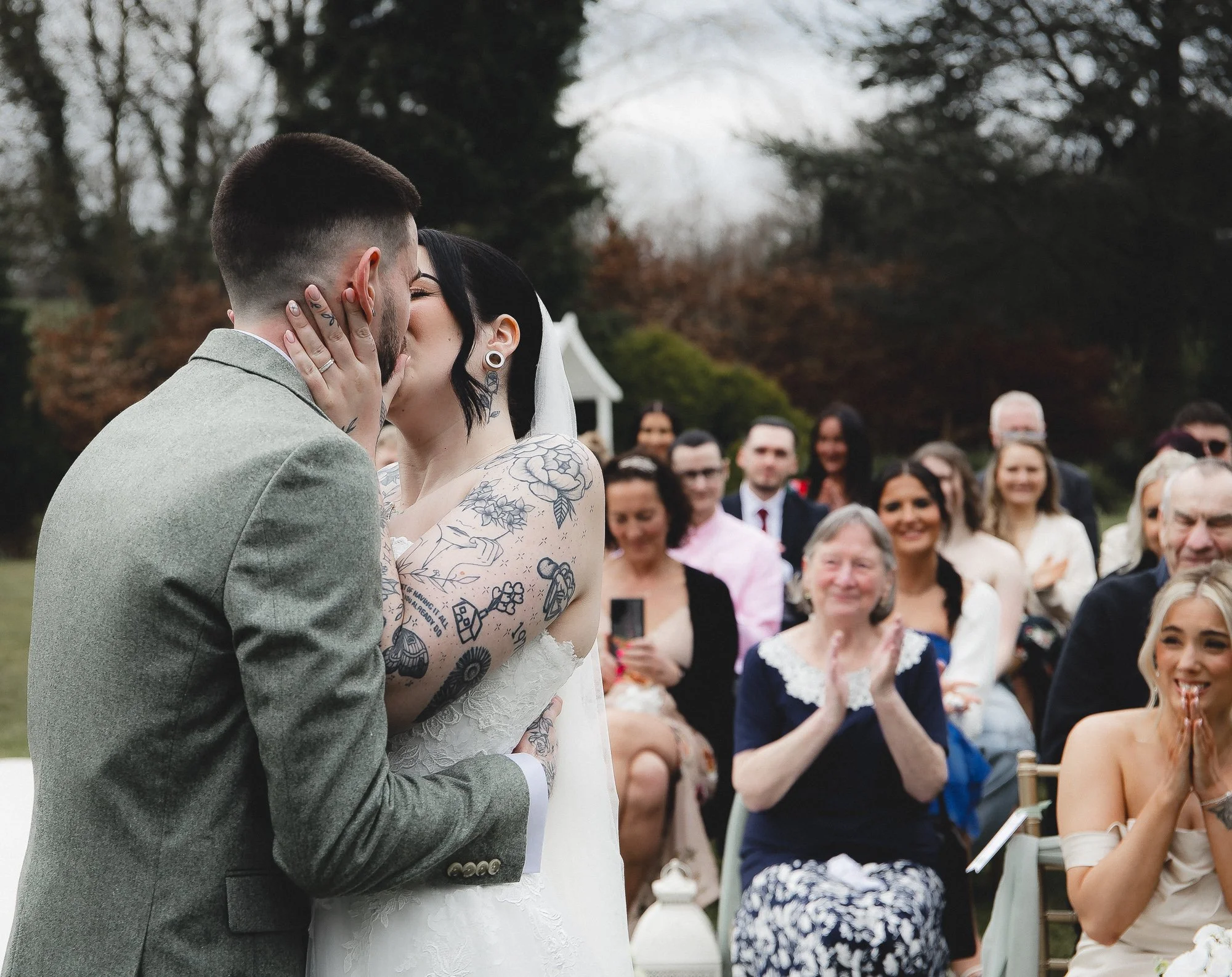 Bride and groom kissing outdoors during wedding ceremony with guests clapping and smiling.