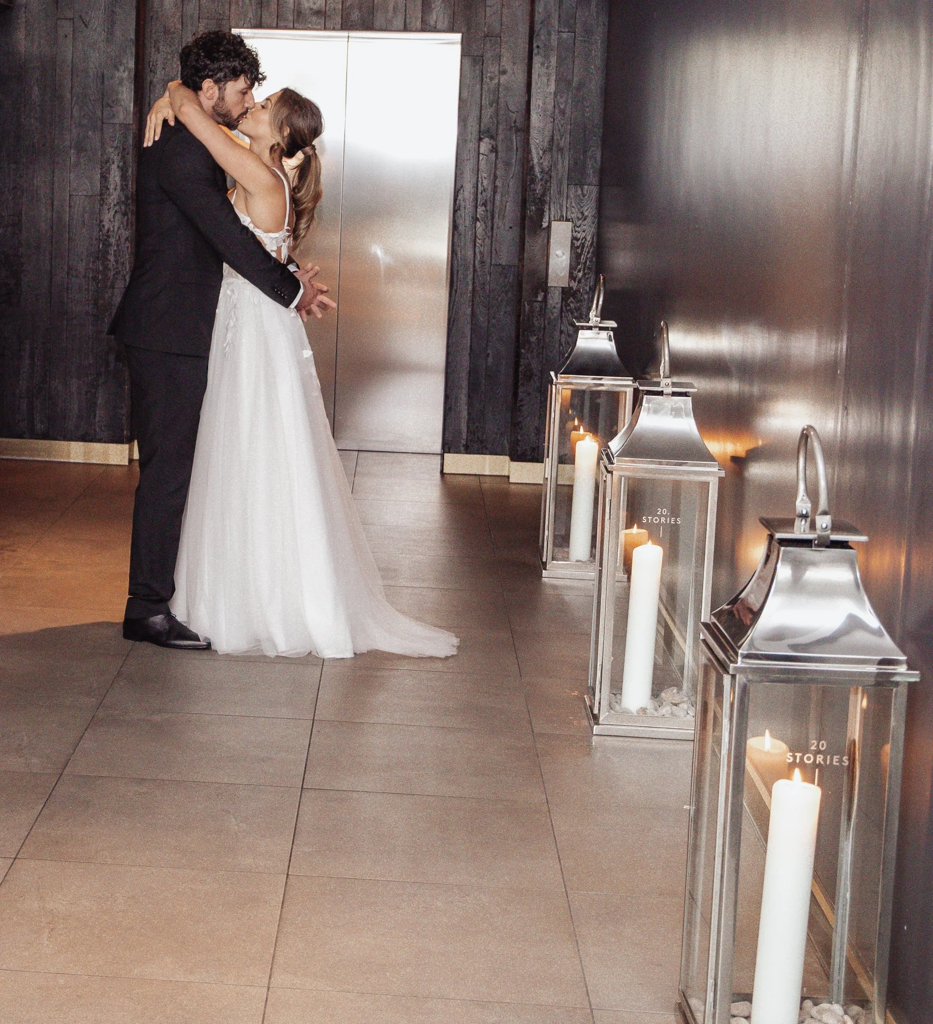 A newlywed couple embracing in a dimly lit hallway with large candle lanterns on the side.