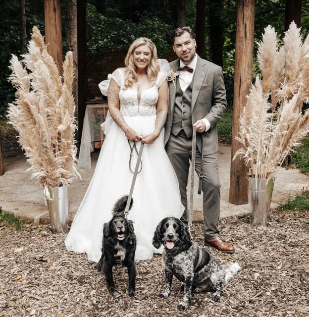A bride and groom stand together in a wooded area, both holding leashes of two dogs. The bride is in a white wedding dress, and the groom is in a grey suit. Pampas grass decor is on either side of them.