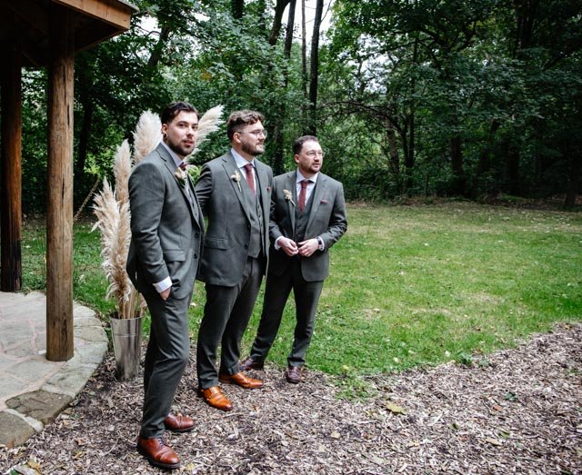 Three men in suits standing outdoors near a wooden structure and tall grass, with a wooded area in the background.