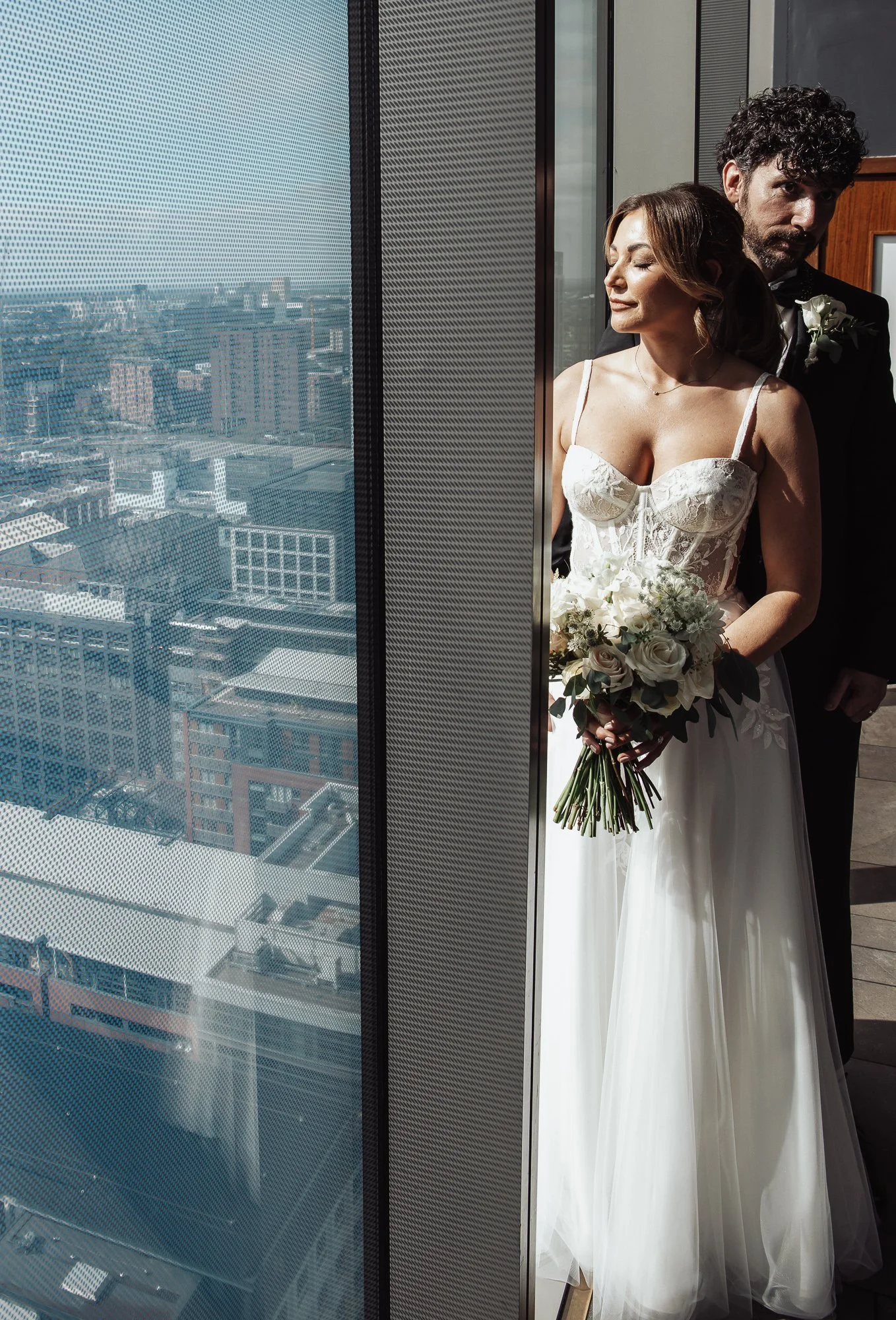 Bride and groom standing by a window with cityscape view, bride holding a bouquet of white flowers.