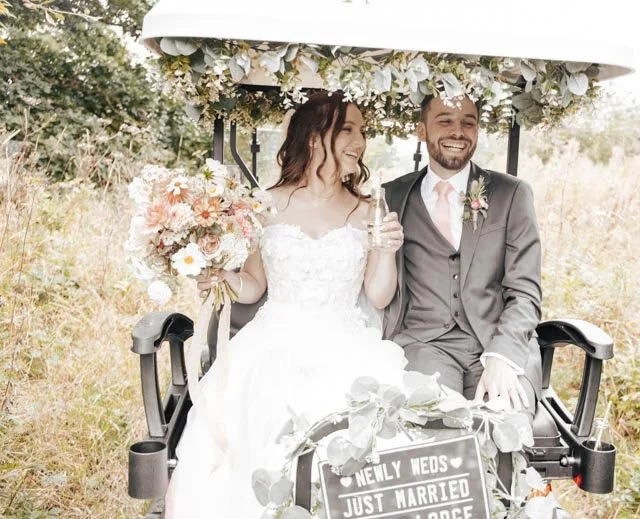 A bride and groom sitting in a decorated golf cart with a "Newly Weds Just Married" sign, surrounded by floral arrangements in a grassy outdoor setting.