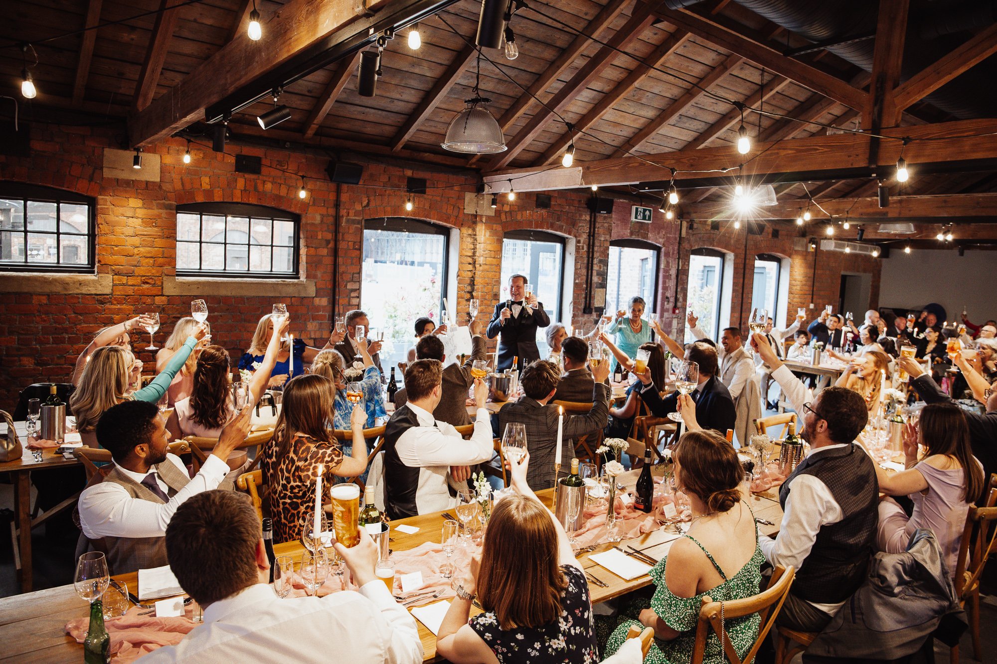 A large gathering of people at a banquet style event in a brick-walled, rustic venue with exposed wooden beams, raising glasses in a toast, with a man in a suit speaking at the front.