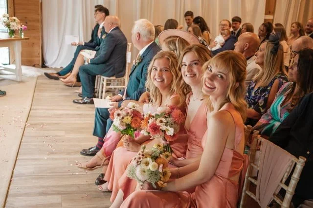 Three bridesmaids in pink dresses holding bouquets, sitting in a row during a wedding ceremony, with other guests seated behind them in a decorated indoor venue.