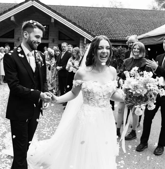 Bride and groom smiling as they walk down the aisle with guests applauding and showering them with confetti.
