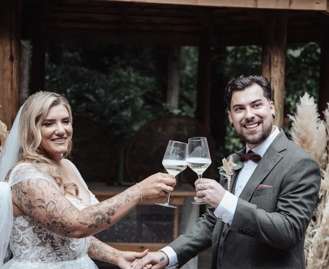 Bride and groom toasting with wine glasses, outdoor setting, wedding attire, smiling, greenery in background.