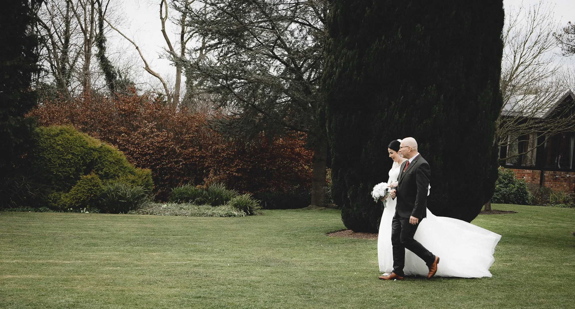 Bride and groom walking in garden, surrounded by trees and bushes, with a grassy lawn.