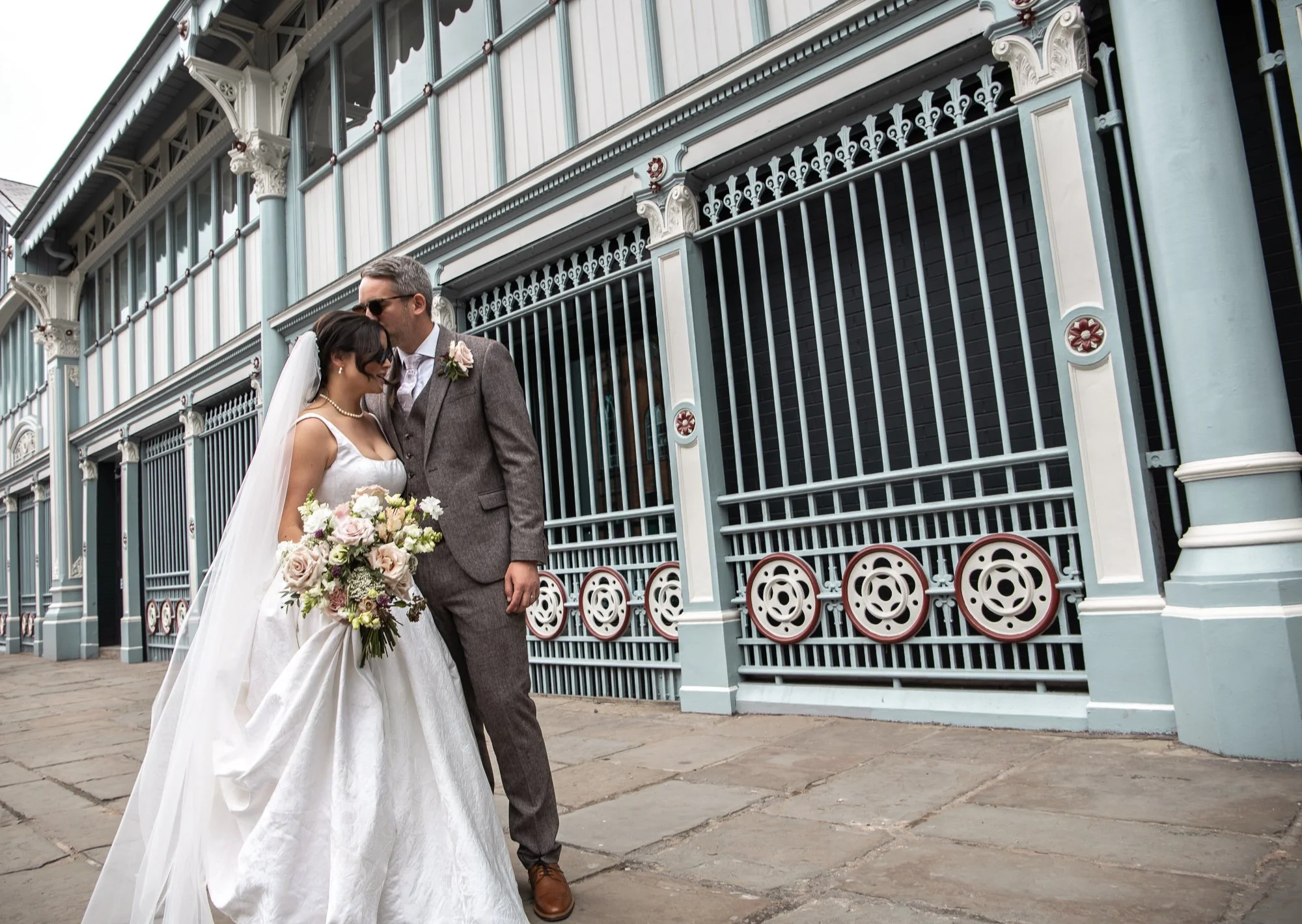 A bride and groom standing close together in front of a decorative metal gate, with the groom kissing the bride on her forehead. The bride is holding a large bouquet of flowers, and both are dressed in wedding attire.