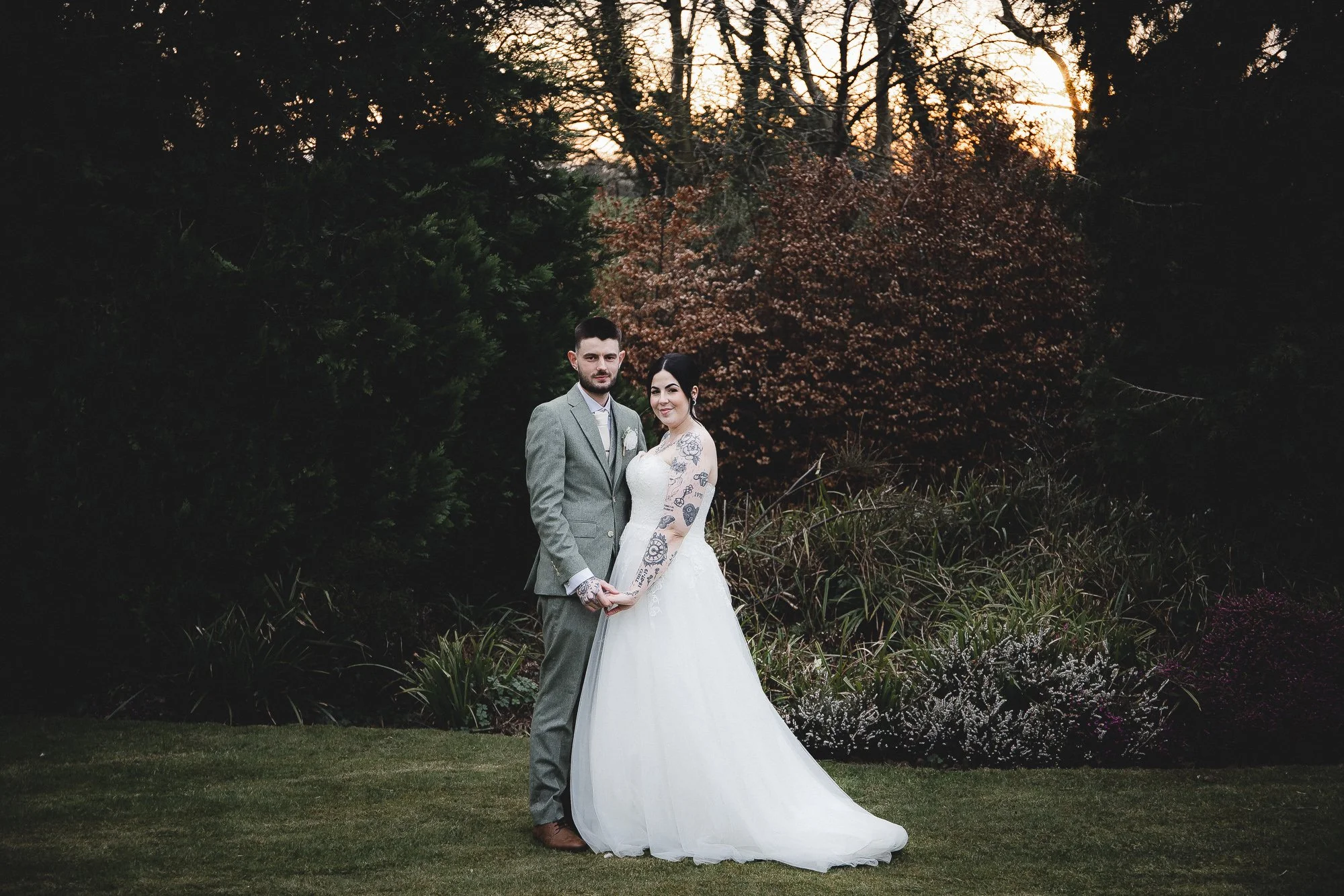 Bride and groom standing outdoors, posing for a wedding photo, with trees and bushes in the background.