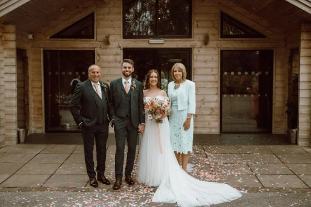 A group of four people in formal attire pose in front of a wooden building at a wedding. The scene includes a bride in a white dress with a long train holding a bouquet, a groom in a suit, and two older adults, one man and one woman. Confetti is scattered on the ground.