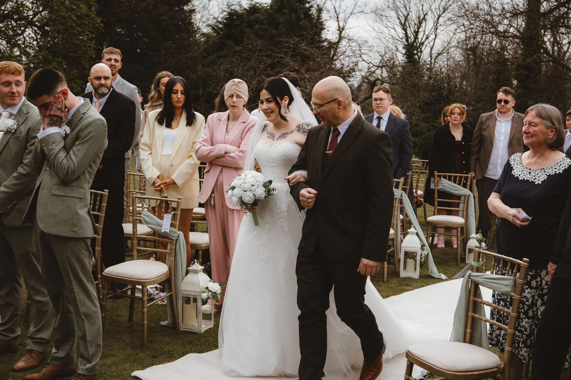 A bride in a white gown holding a bouquet, walking down an outdoor aisle with an older man. Guests watch, seated and standing on either side, with trees in the background.