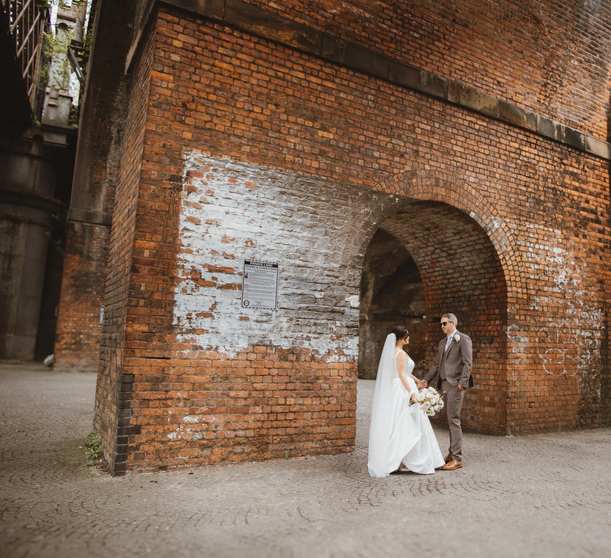 A bride and groom standing under a large brick archway, holding hands, with the bride holding a bouquet of flowers, in an urban setting with brick walls and graffiti.