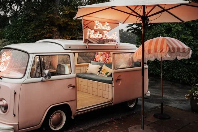 Retro van converted into a photo booth with open sliding door, neon sign "Birdie Photo Booth," inside seating, and an umbrella nearby.