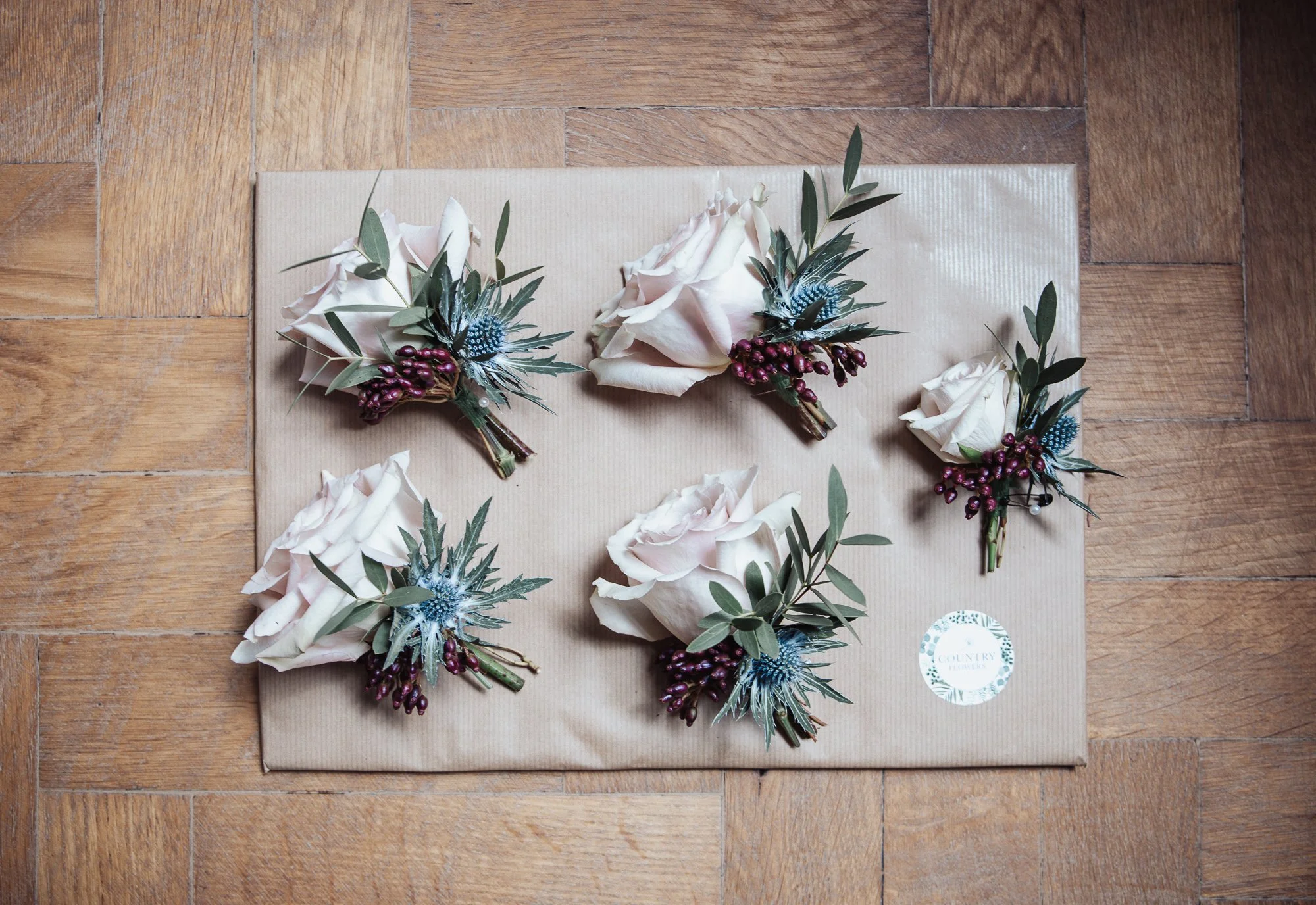 Five floral boutonnières with white roses, blue thistles, and purple berries arranged on a beige gift box on a wooden floor.