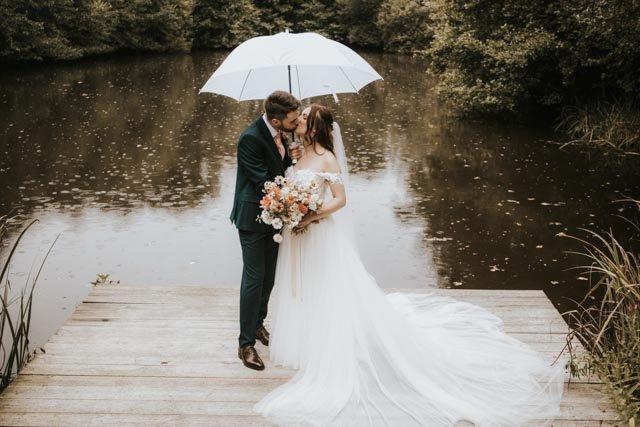 Bride and groom kissing under a white umbrella on a wooden dock by a lake.