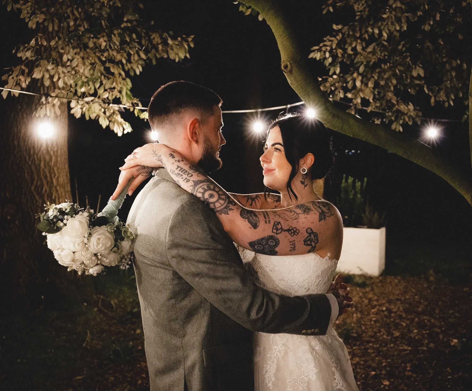 A bride and groom embracing outdoors at night, with string lights overhead. The bride, wearing a white dress and holding a bouquet, has tattoos on her arms. The groom is in a suit.