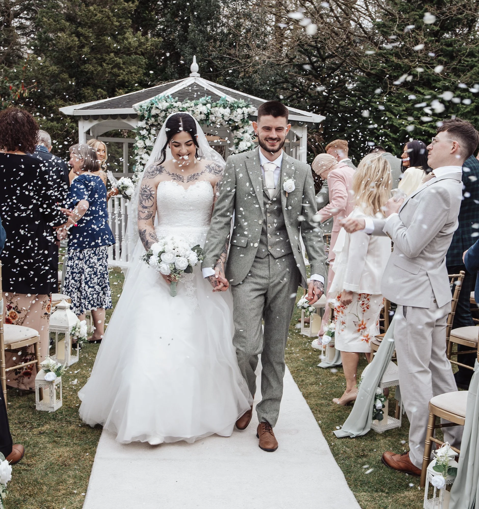 Bride and groom walking down the aisle at an outdoor wedding ceremony, surrounded by guests throwing confetti, with a gazebo decorated with flowers in the background.
