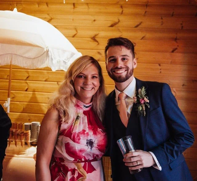Two people smiling, one wearing a floral dress and the other in a suit with a peach tie, standing indoors in front of wooden paneling and a white umbrella.