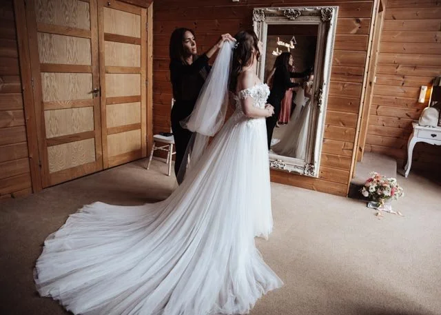 Bride getting ready in a wooden room with a large mirror, attended by a woman adjusting her veil. The bride is wearing a white wedding dress with a long train. A bouquet of flowers is on a table nearby.