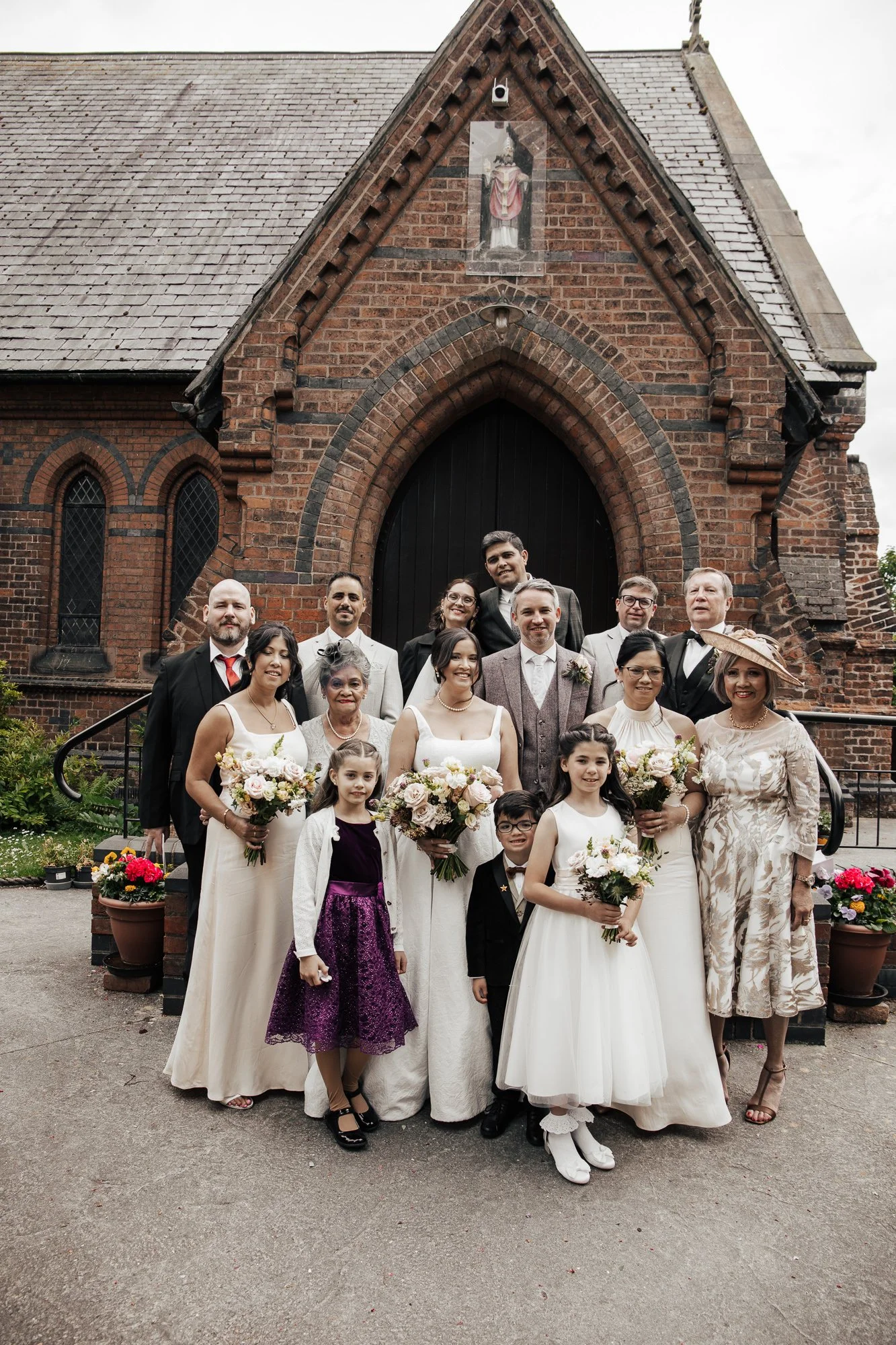 A wedding group photo with the bride, groom, family, and friends standing in front of a brick church with stained glass windows and religious statues. The bride and some women hold bouquets, and the children are dressed in formal attire.