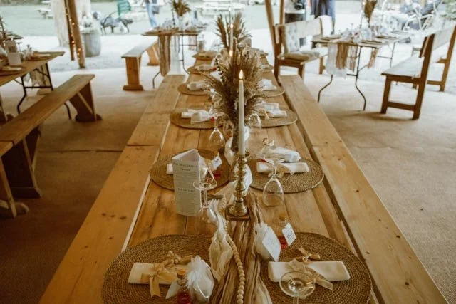 Rustic table setting with wooden tables, woven placemats, white napkins, wine glasses, and a lit candle centerpiece, designed for an outdoor event or wedding reception.