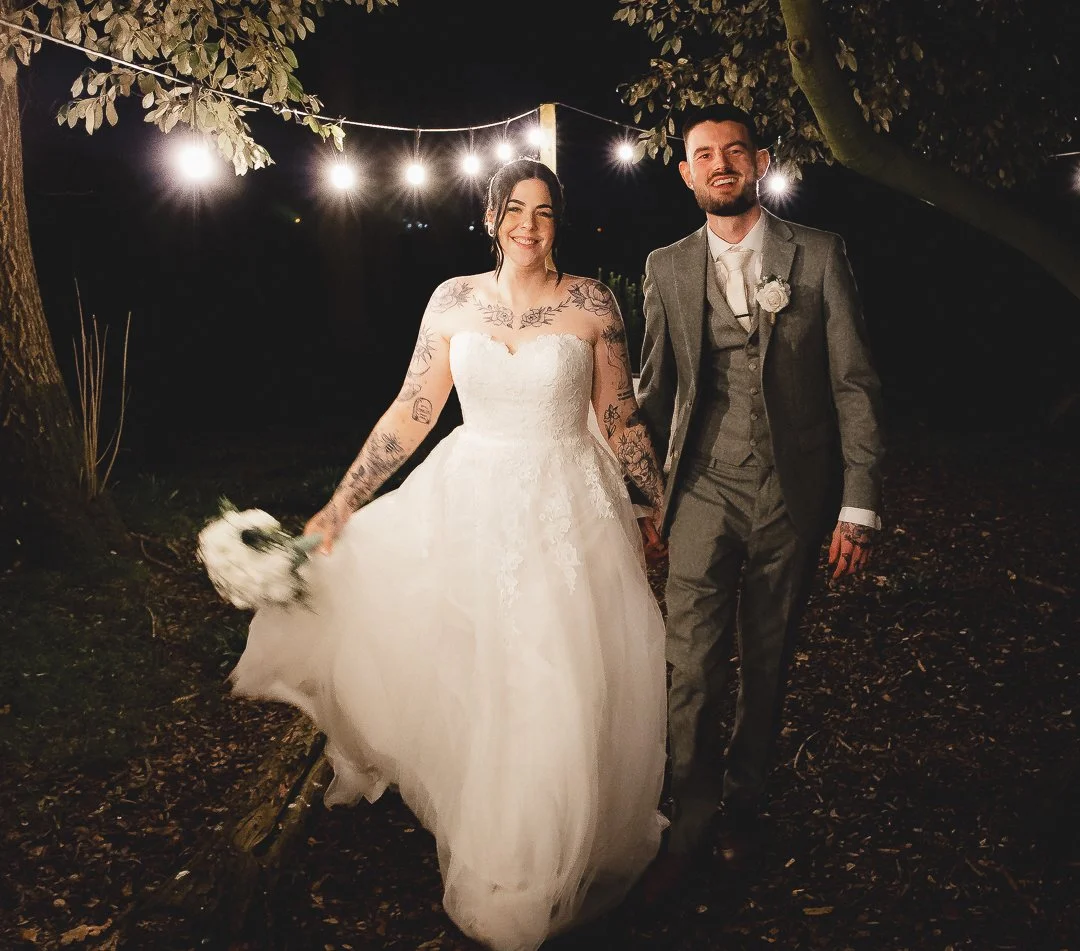Bride and groom smiling and holding hands at an outdoor wedding under string lights, bride in a white dress holding a bouquet, groom in a gray suit.