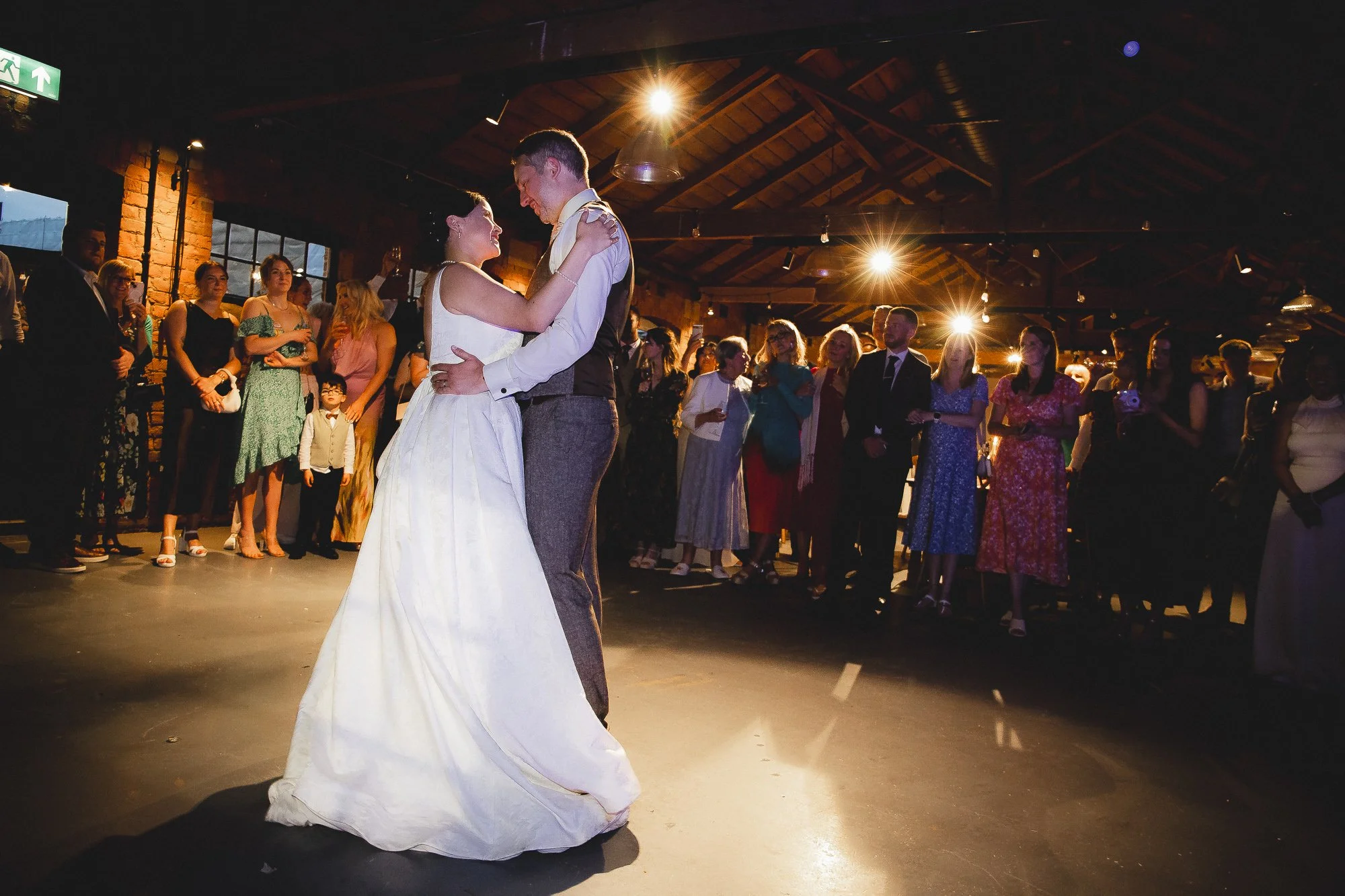 A bride and groom are dancing at their wedding reception with guests watching in a dimly lit venue with exposed brick walls and wooden ceiling beams.