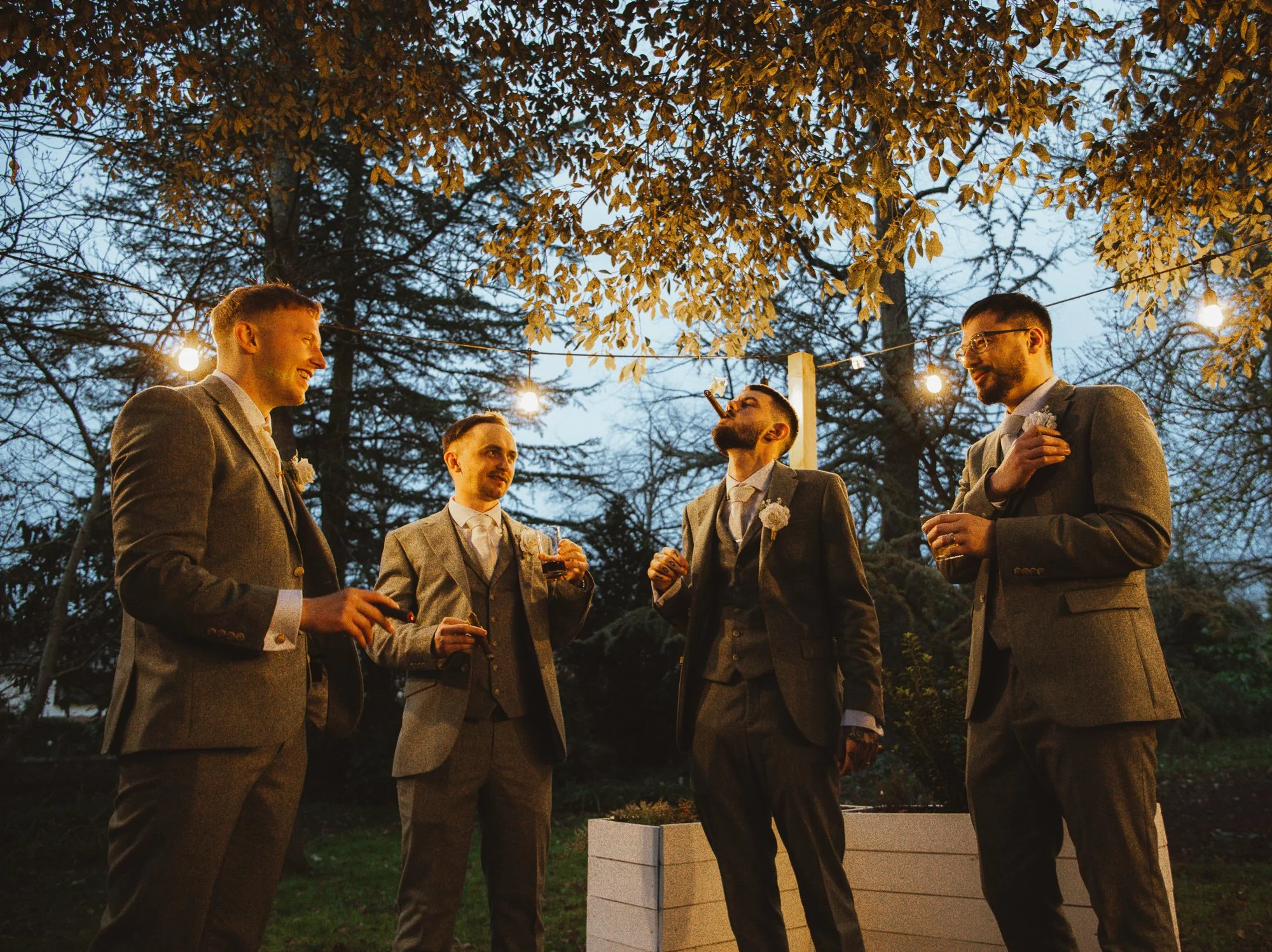 Four men in suits enjoying cigars and drinks outdoors at dusk.