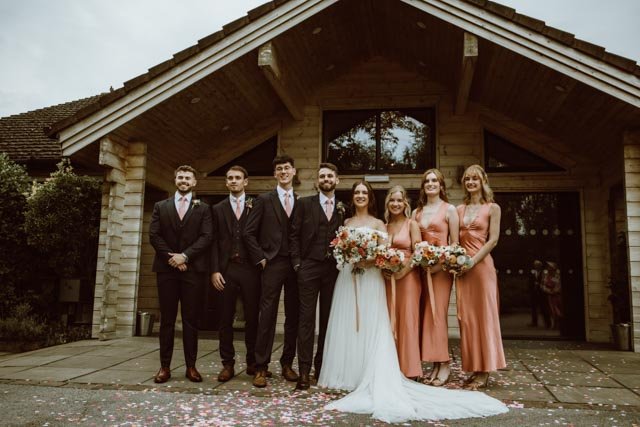 Wedding party posing in front of a rustic wooden building, with the bride in a white dress holding a bouquet of flowers, surrounded by bridesmaids in pink dresses and groomsmen in dark suits.