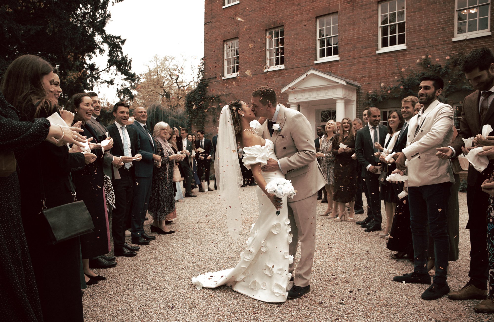 A bride and groom kiss amidst wedding guests outside a brick building, with guests throwing flower petals in celebration.