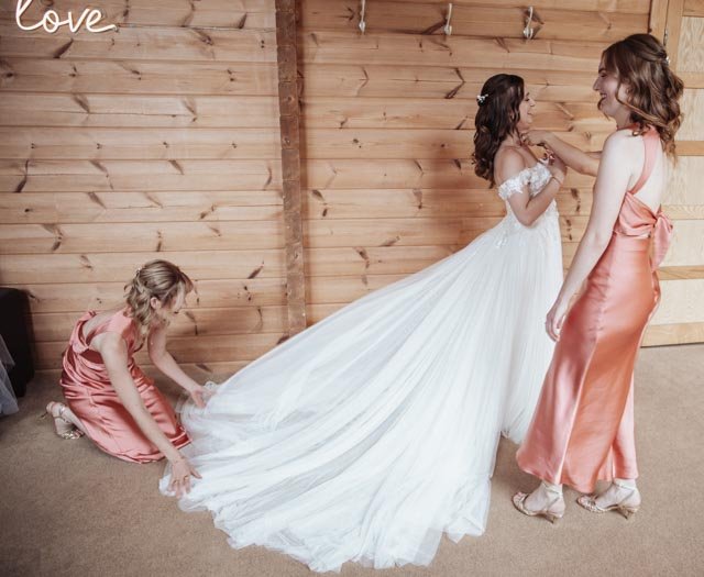 Bride in a white wedding dress with bridesmaids in peach dresses adjusting her gown in a rustic wooden room.