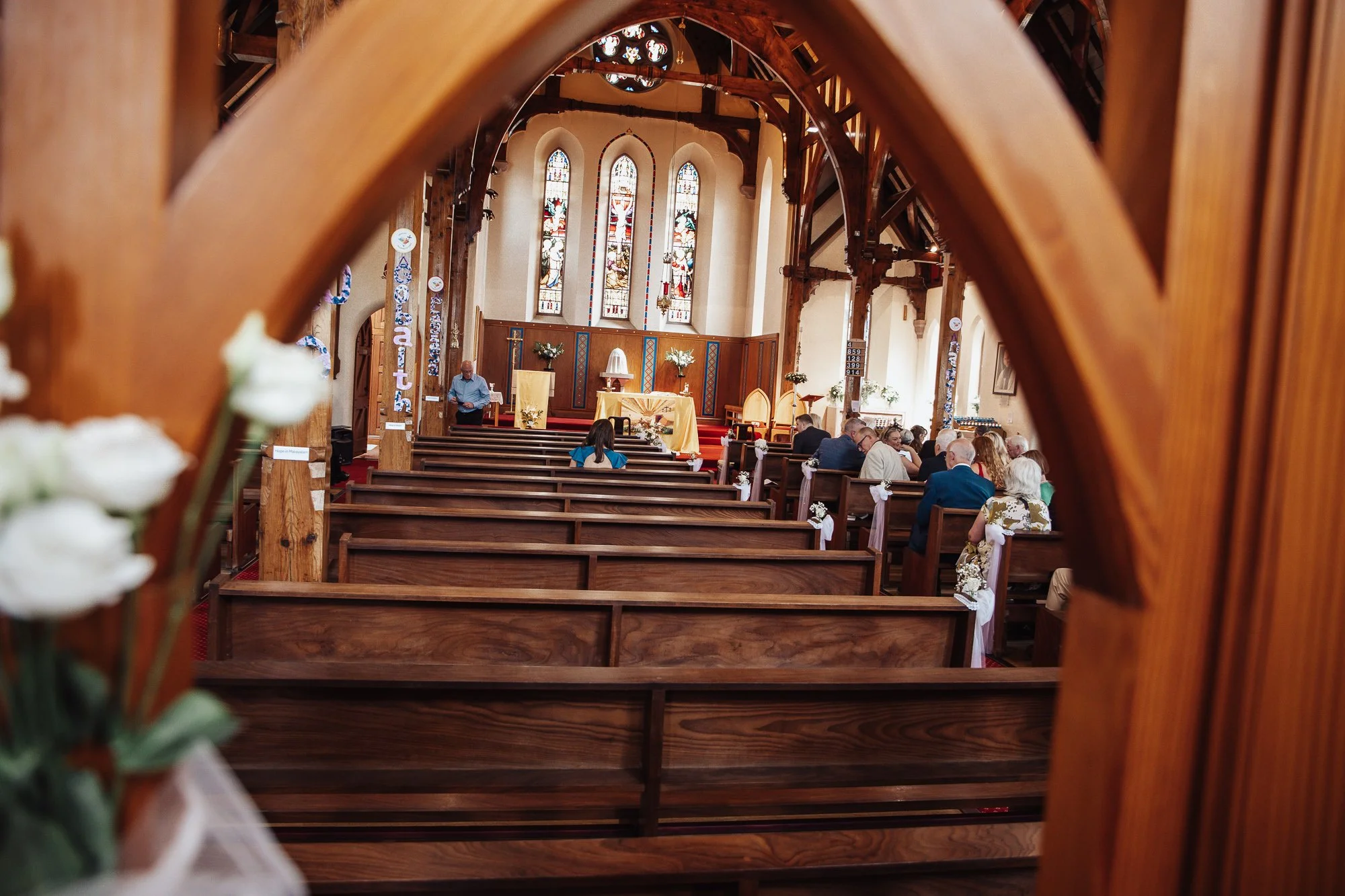 Inside a church during a wedding ceremony, with guests sitting in pews, decorated with white flowers. The altar has candles and flowers, and stained glass windows are in the background.