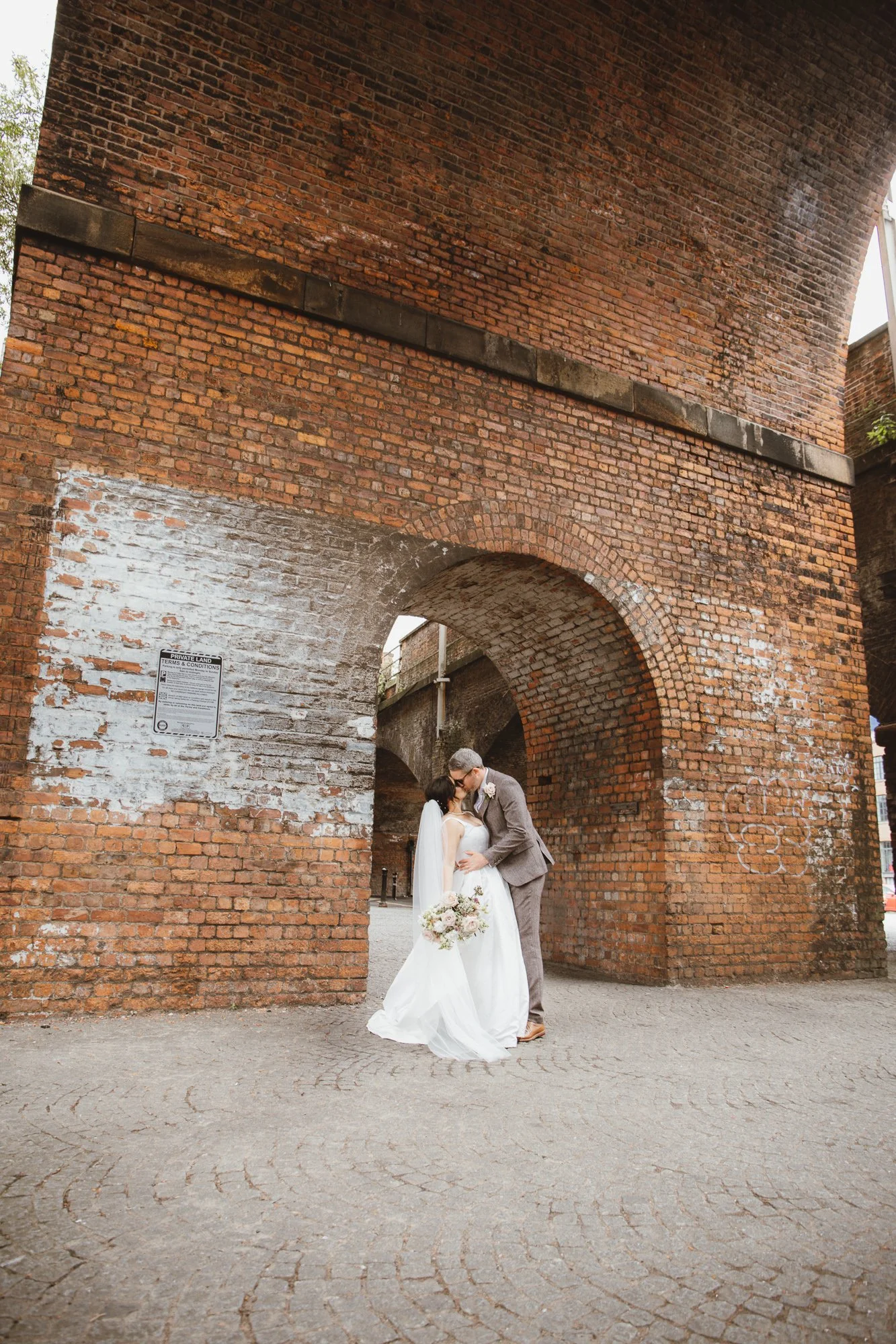 A bride and groom sharing a kiss under a large brick archway, with the groom dressed in a gray suit and the bride holding a bouquet of flowers.