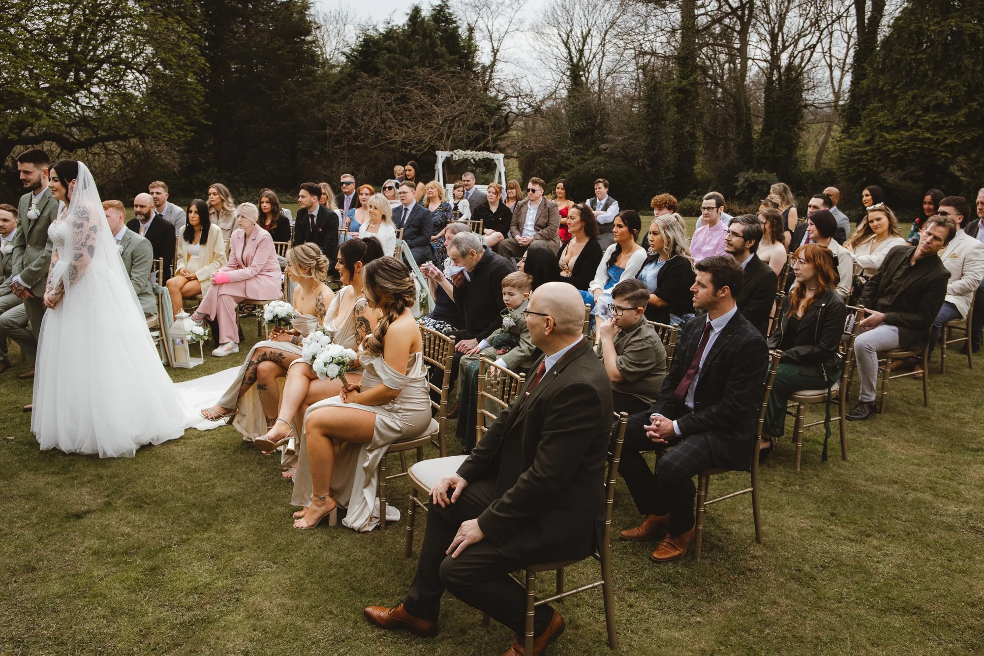 Outdoor wedding ceremony with guests seated, bride and groom standing in the foreground, and trees in the background.