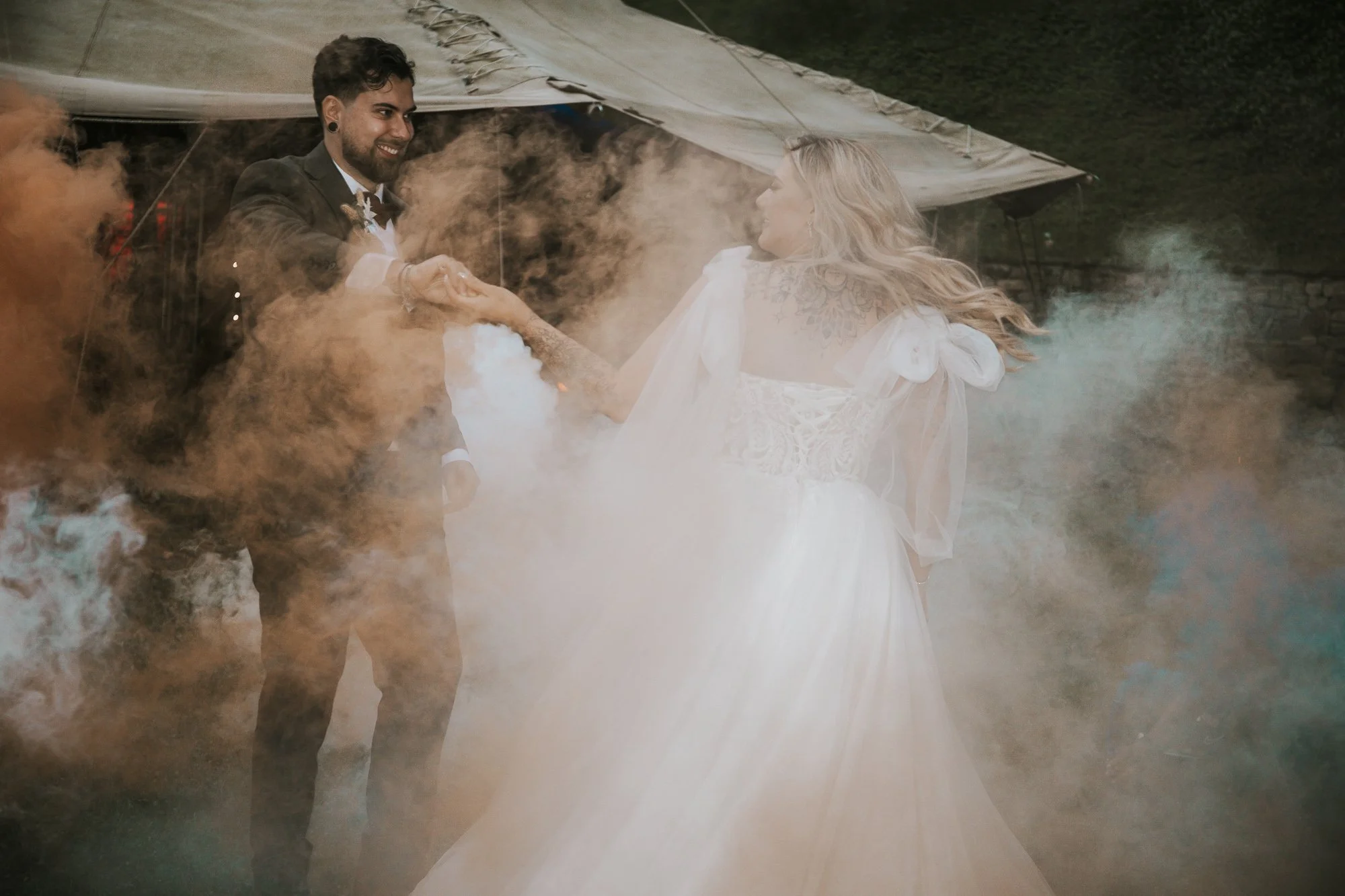 A couple dancing surrounded by colorful smoke, with the woman in a white dress and the man in a suit, outdoors under a tent.