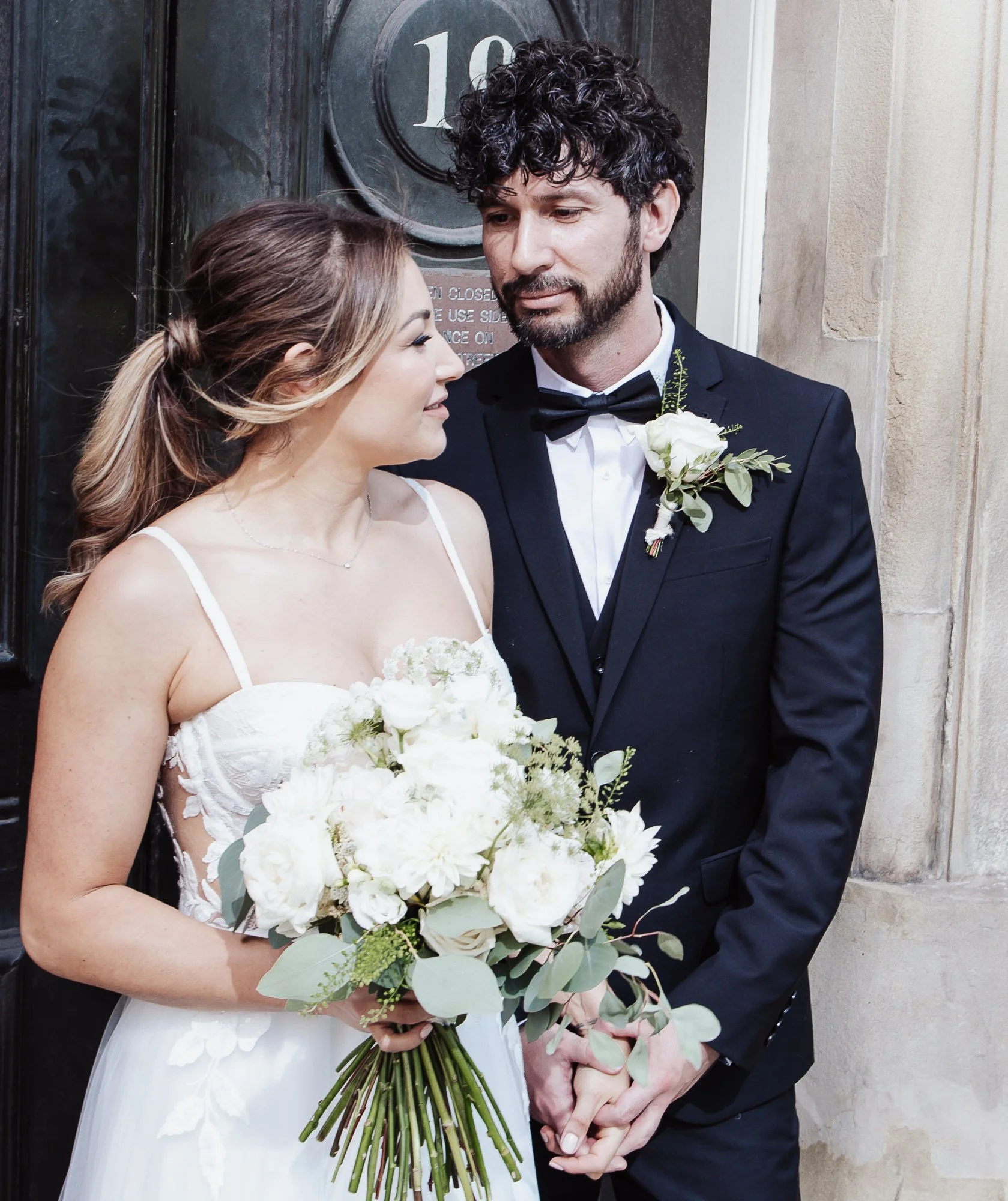 Bride and groom holding hands, the bride is holding a bouquet of white flowers, standing in front of a door with the number 16.