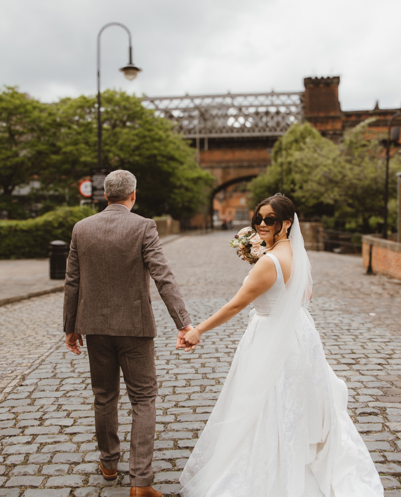 A newlywed couple holding hands on a cobblestone street, with the bride wearing a white wedding dress and sunglasses, and the groom in a gray suit, under an overcast sky.