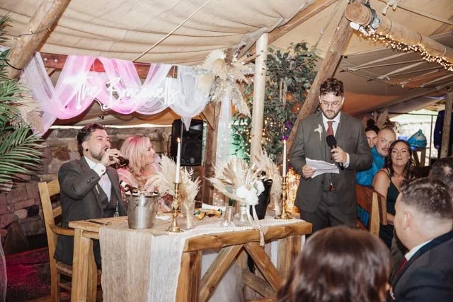 A wedding reception scene with a couple sitting at a decorated wooden table while a man in a suit speaks into a microphone. The setting is adorned with lights, foliage, and rustic decorations. Guests are sitting and observing the speech.