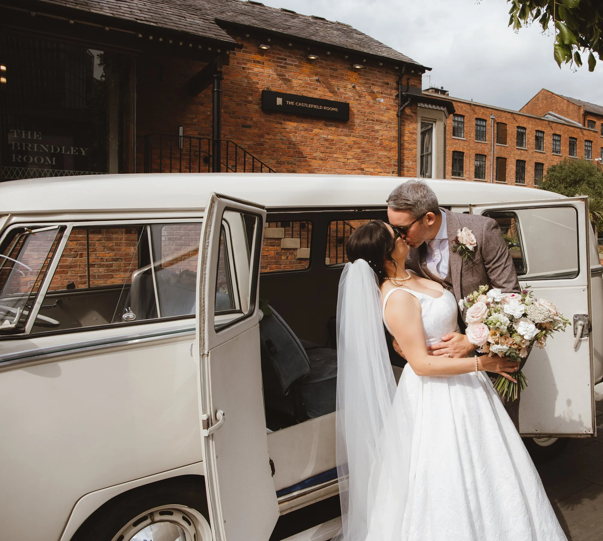 A bride and groom sharing a kiss outside a vintage white car, with the bride holding a bouquet of flowers and wearing a white wedding dress with a veil, in front of a brick building.