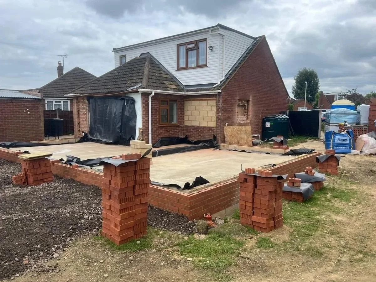 Construction site with brick foundation and partially built house, construction materials, and tools, in a residential neighborhood.