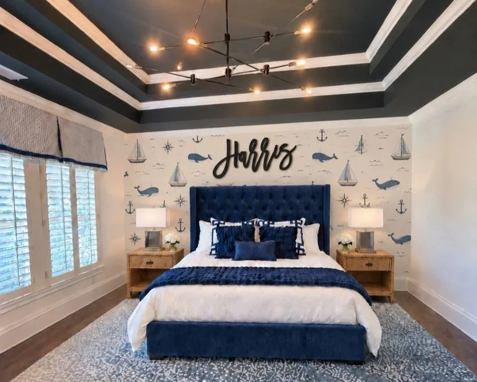 Nautical boys bedroom with navy tufted bed, sailboat and whale wallpaper, wood name sign, and dark tray ceiling with chandelier.