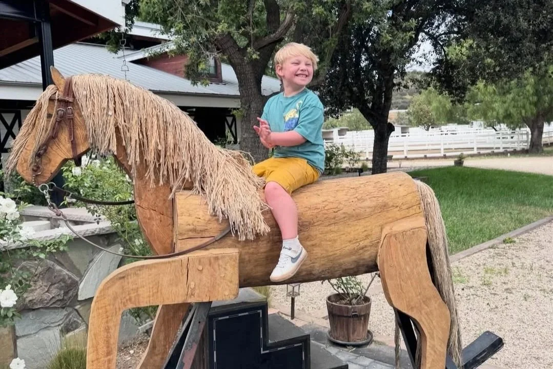 Young boy laughing while riding a large wooden horse statue outdoors.
