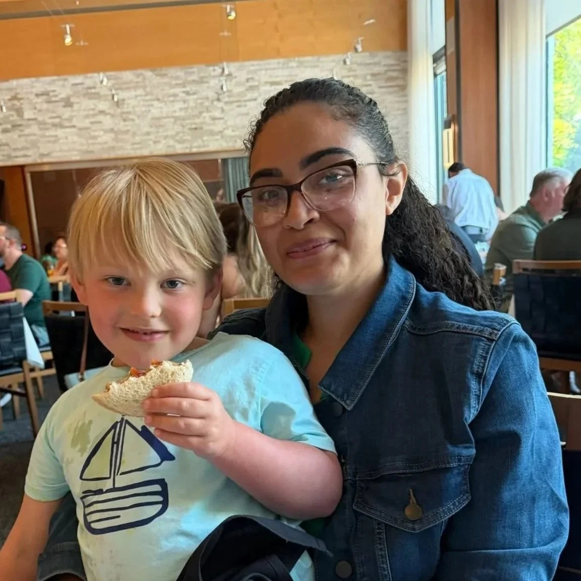 Woman holding a young boy eating a sandwich at a restaurant table.