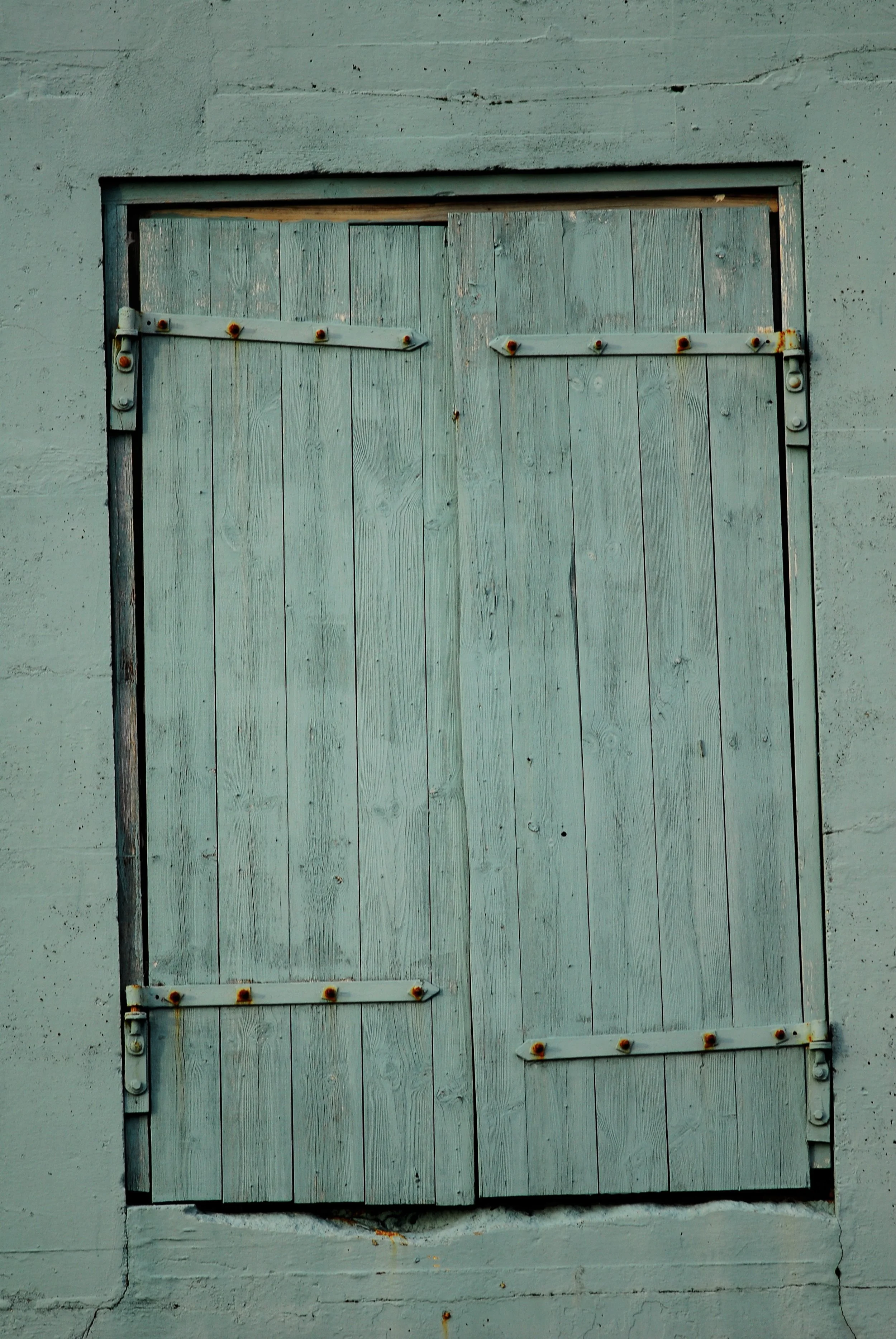 Weathered wooden window shutters painted in faded blue, mounted on a gray concrete wall with rusted hardware.