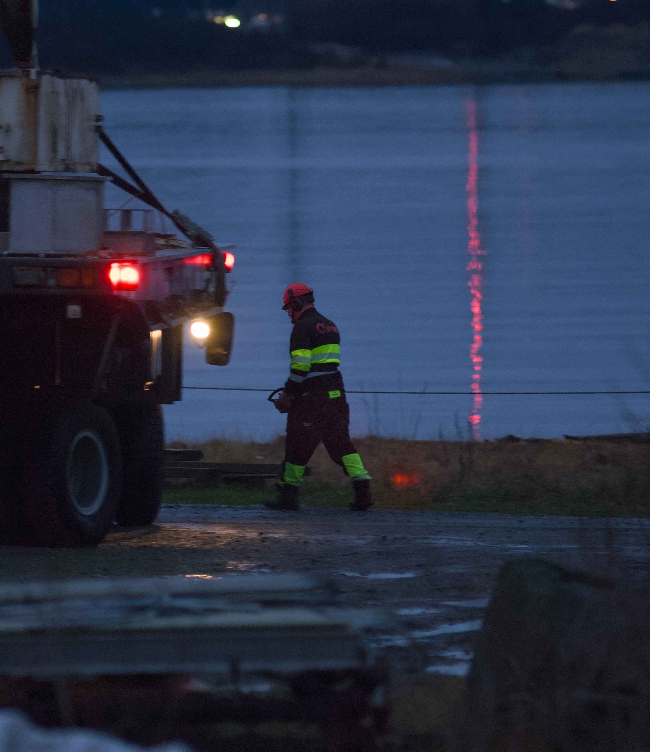 A firefighter in protective gear and a red helmet stands next to a fire truck near water during dusk or early evening.